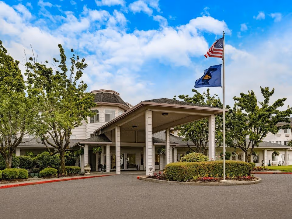 Exterior view of Parkrose Chateau Gracious Retirement Living facility showing the main entrance with a covered driveway, surrounded by trees and bushes. Two flags, including the American flag, are flying on a flagpole near the entrance under a partly cloudy blue sky.