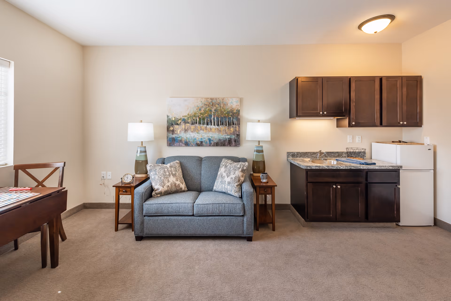 Cozy living area with a loveseat between two side tables and lamps, wall art above, and a small kitchenette with dark cabinets and a mini-fridge to the right.