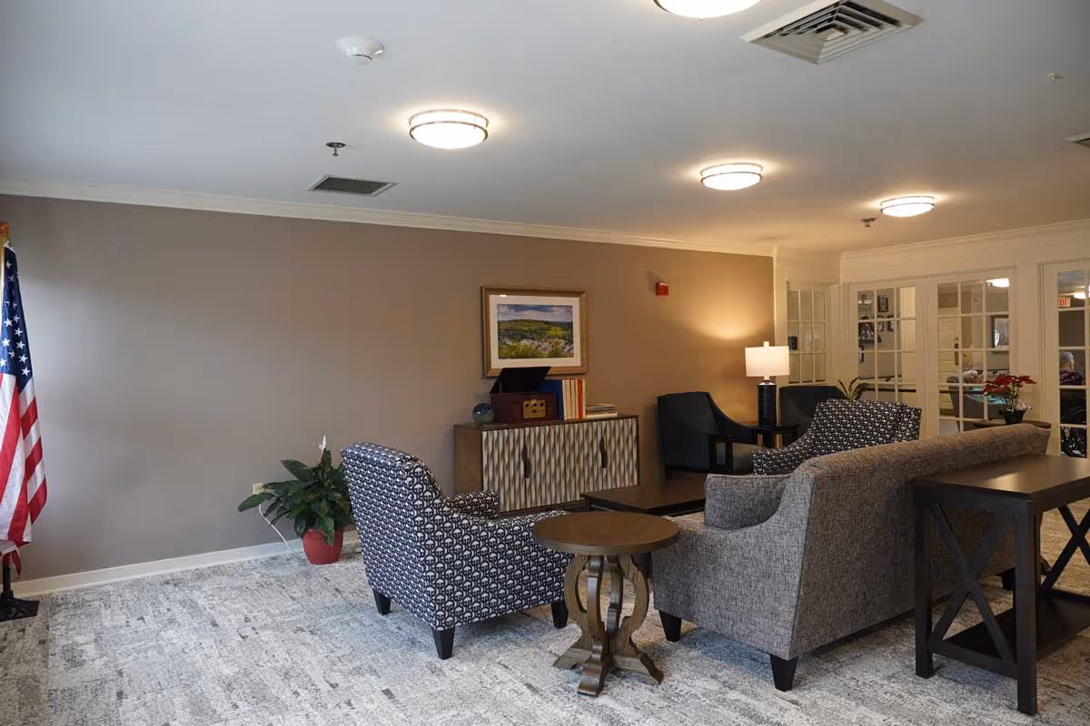 A cozy living room area with patterned armchairs, a gray sofa, a wooden side table, and a cabinet against a beige wall. There is a framed landscape picture above the cabinet, a potted plant, an American flag, and a lamp on a side table. The room has carpeted flooring and ceiling lights.
