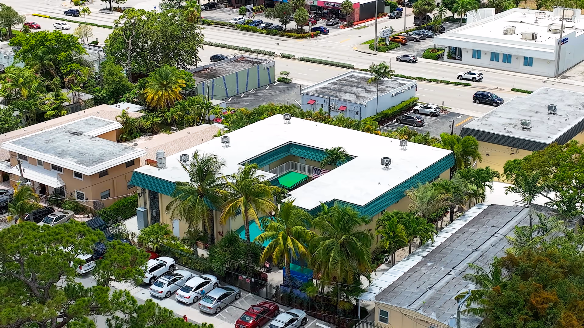 Aerial view of a senior living facility named Villa Rio Vista, showing a U-shaped building with a white roof and green trim surrounded by palm trees and other greenery. There is a parking lot with several cars parked, and nearby streets with additional buildings and vehicles are visible.