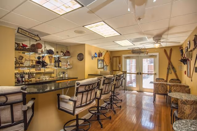 Interior view of a senior living community lounge area featuring a bar with cushioned swivel bar stools, a mirrored back wall with shelves holding bottles and glassware, and a seating area with small tables and chairs. The room has wooden floors, yellow walls decorated with vintage items including skis, a dartboard, and framed pictures, and double glass doors letting in natural light.