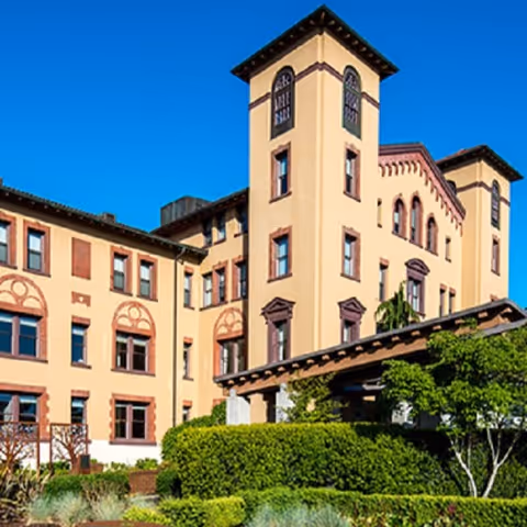 Tall beige senior living building with towers, arched windows, and manicured landscaping.