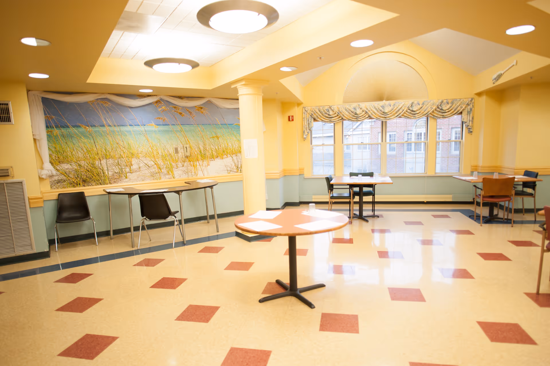 A bright room with yellow walls and a tiled floor featuring red and beige squares. There are several tables and chairs arranged around the room. A large mural of a beach scene with sand dunes and sea oats decorates one wall, and a large window with a decorative valance lets in natural light. The ceiling has recessed lighting and circular light fixtures.
