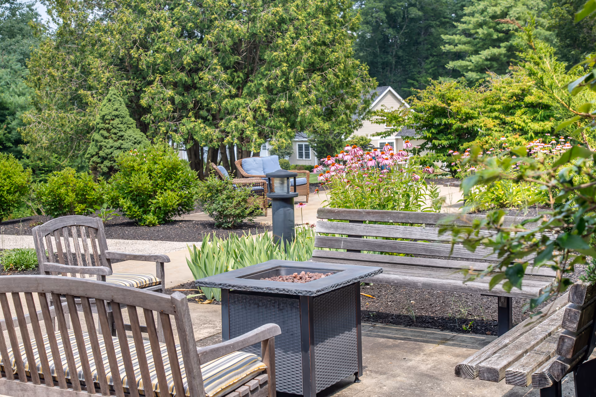 Outdoor seating area with wooden benches and chairs around a square fire pit table, surrounded by green bushes, pink flowers, and trees with a house visible in the background.