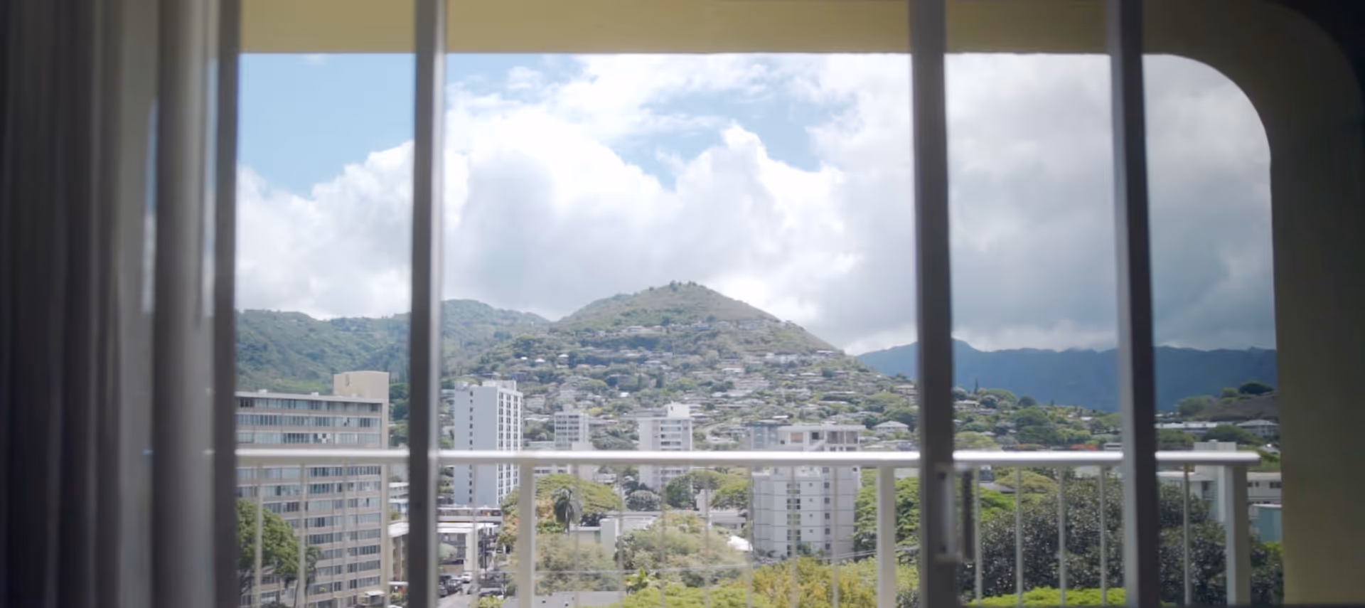 View through a sliding glass door with vertical bars, looking out onto a balcony railing and a cityscape with buildings and green hills under a partly cloudy sky.