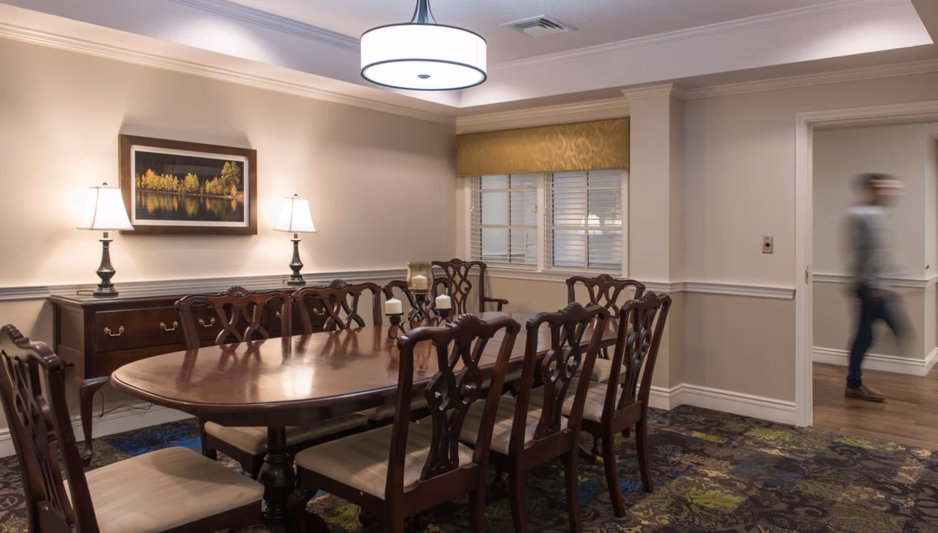 A formal dining room with a long wooden table surrounded by chairs, a sideboard with lamps and wall art, and a blurred person walking through a doorway.