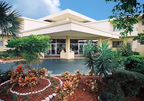 Front exterior view of Oasis at Margate Assisted Living Residence showing the entrance with large glass doors, surrounded by landscaped garden beds with various shrubs and plants under a partly cloudy sky.