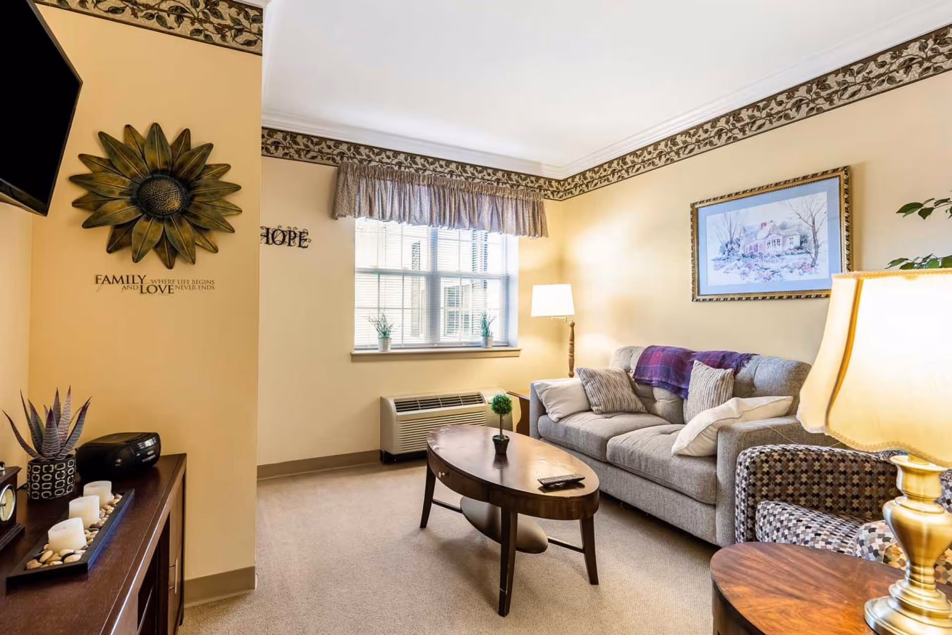 A cozy living room with a gray sofa adorned with pillows and a purple throw blanket, a patterned armchair, a wooden coffee table with a small plant and remote control, a floor lamp, and a window with blinds and a valance. The walls are painted light yellow with a floral border near the ceiling. Decorative items include a sunflower wall art, a framed picture of a house, and a sideboard with candles and a small plant.