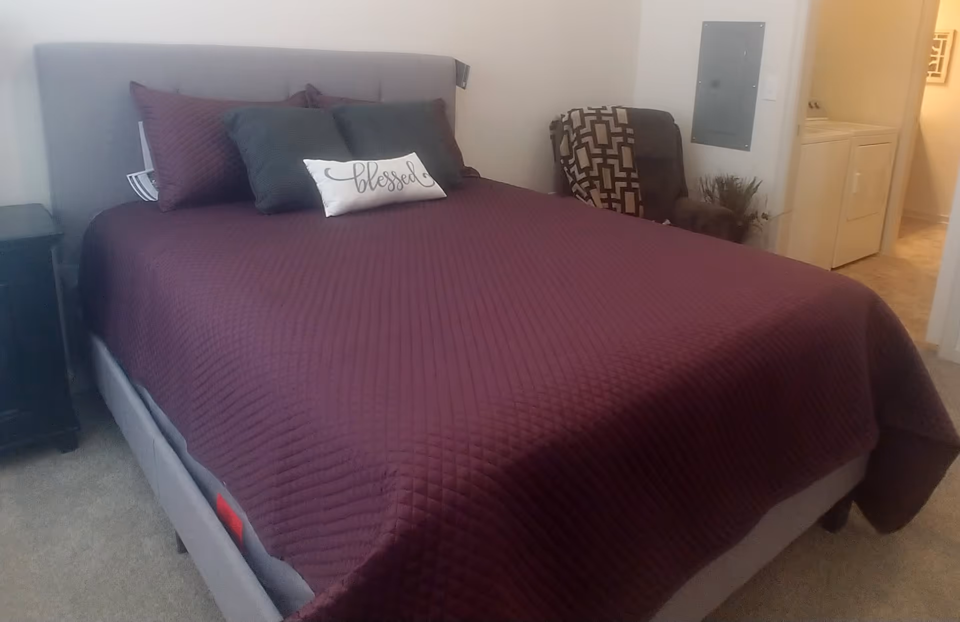 A neatly made bed with a purple quilt and decorative pillows (one reading "blessed") in a bedroom with a chair and a laundry alcove.