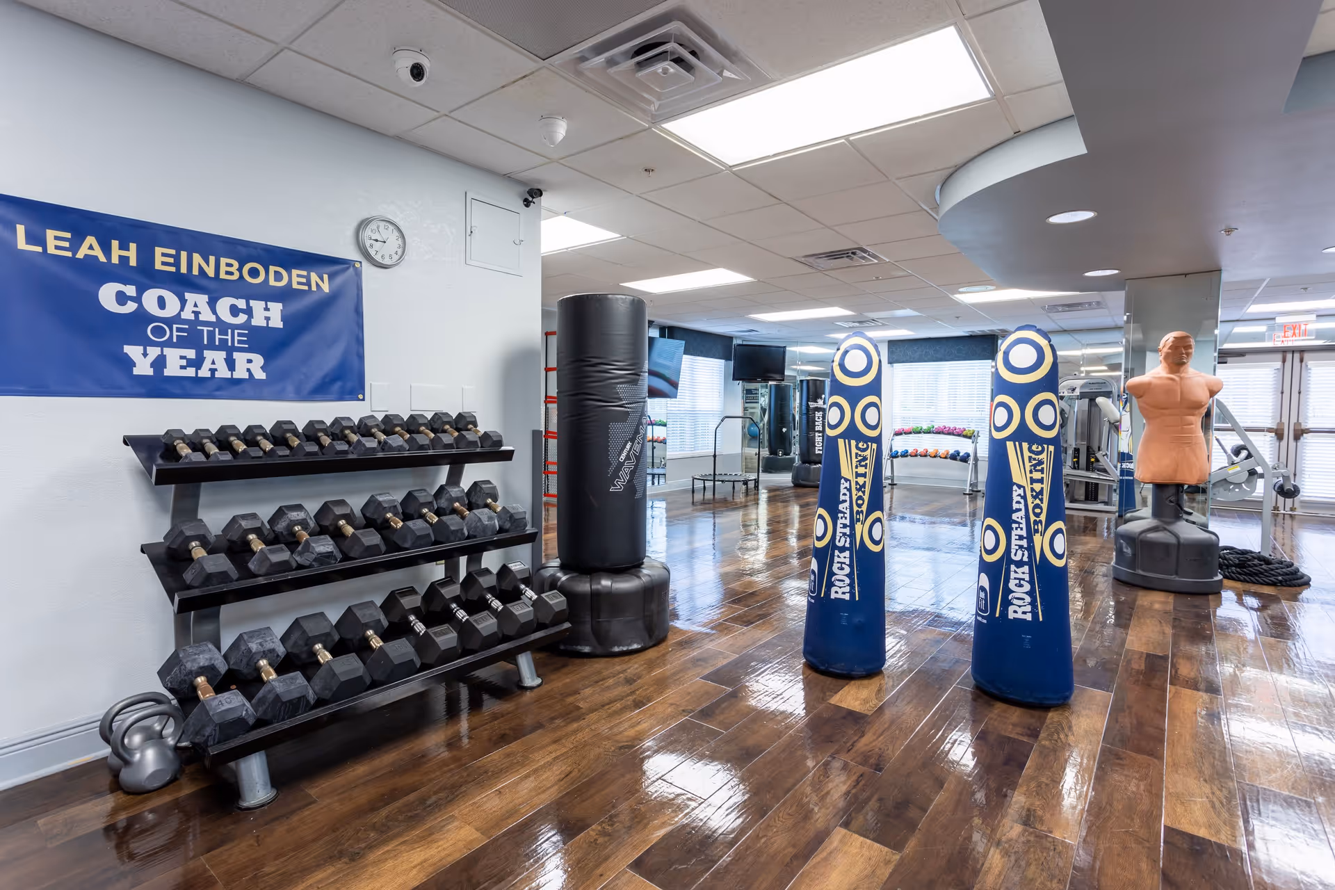 Fitness room with a dumbbell rack, free weights, standing punching bags and a training dummy on a polished wood floor.