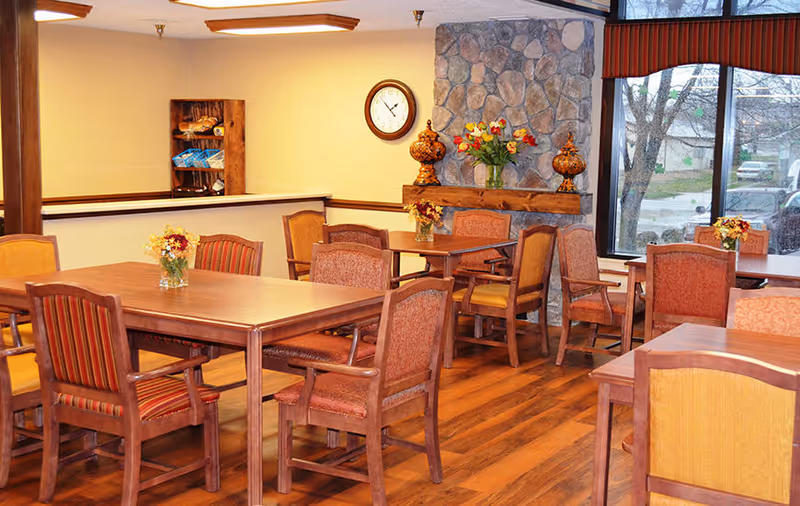 Wooden dining room with multiple tables and upholstered chairs, floral centerpieces, a stone fireplace wall, and large windows.
