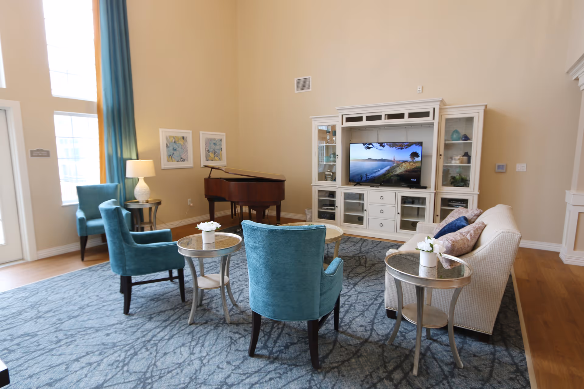 A bright and spacious living room with high ceilings, featuring a white entertainment center with a flat-screen TV displaying a scenic view. The room has a beige sofa with decorative pillows, three teal upholstered chairs, and three round side tables with small white flower arrangements. A grand piano is positioned in the corner near two framed floral artworks on the wall. Large windows with teal curtains allow natural light to fill the space, which has a blue patterned area rug and wooden flooring.