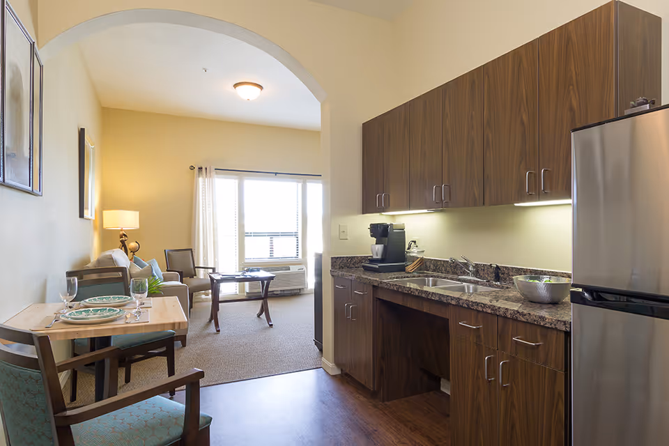 Interior view of a senior living apartment featuring a small kitchen area with wooden cabinets, a granite countertop, a coffee maker, and a stainless steel refrigerator. Adjacent to the kitchen is a dining table set for two with plates and glasses. Beyond the dining area is a living room with a sofa, armchair, coffee table, lamp, and large window with curtains allowing natural light to fill the space.
