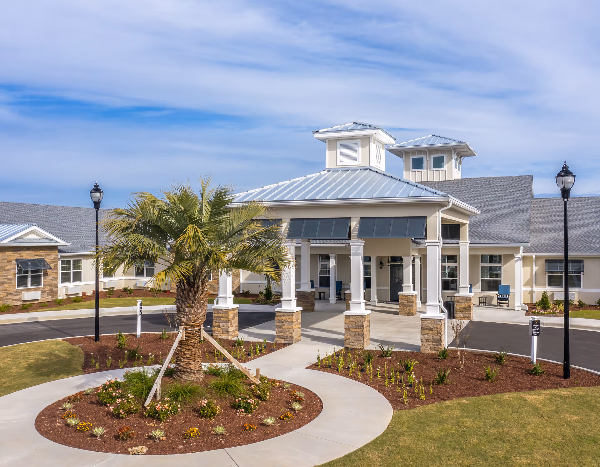 Front entrance of a single-story senior living facility with a covered porte-cochere, landscaped circular driveway and a palm tree.