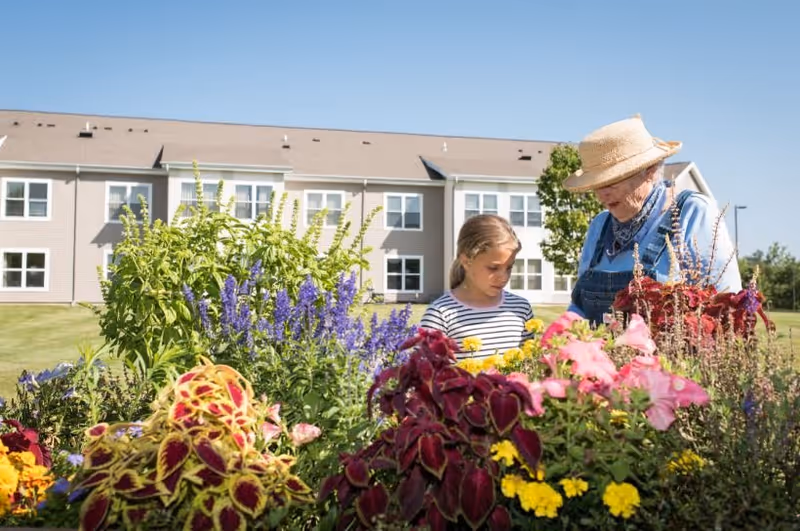 An elderly woman wearing a straw hat and denim overalls is gardening with a young girl in a colorful flower garden. Behind them is a two-story residential building under a clear blue sky.