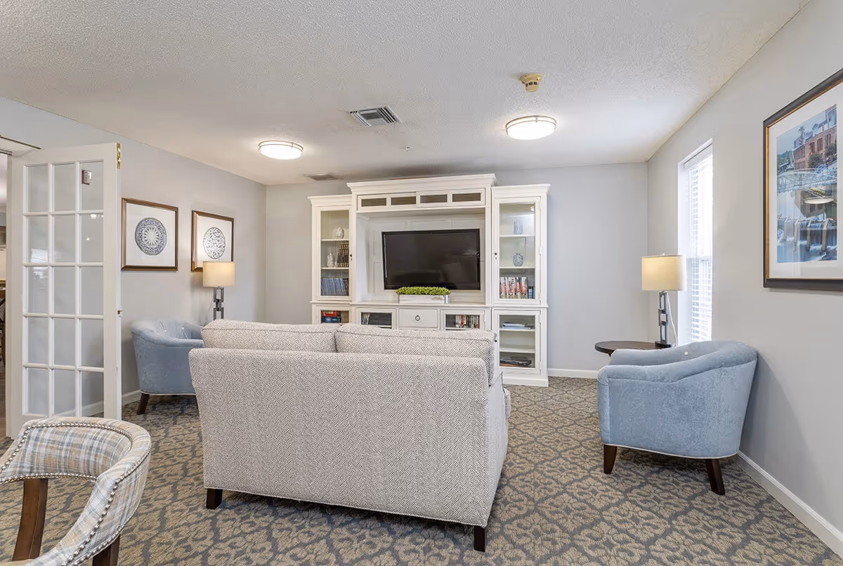 A cozy living room with a gray patterned carpet, a light gray sofa facing a white entertainment center with a flat-screen TV. Two light blue armchairs are positioned on either side of the room, with side tables and lamps next to them. The walls are light gray with framed artwork and a window letting in natural light.