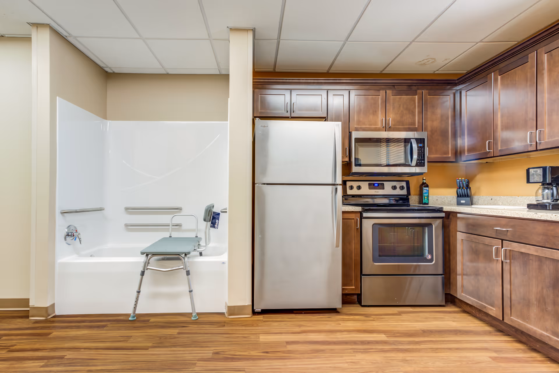 A combined space featuring a white bathtub with grab bars and a shower chair on the left, adjacent to a kitchen area with stainless steel refrigerator, stove, microwave, wooden cabinets, and a coffee maker on the right. The floor is wood laminate and the walls are beige.