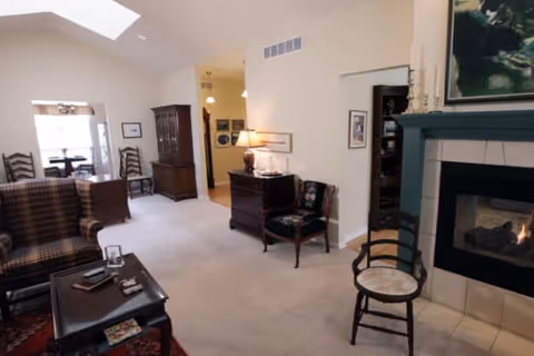 A cozy living room area with a fireplace on the right side, a cushioned chair with floral upholstery, a wooden chair with a woven seat, and a dark wooden coffee table with items on it. The room has beige walls and carpet, a skylight, and a dining area visible in the background with wooden chairs and a table near a window.