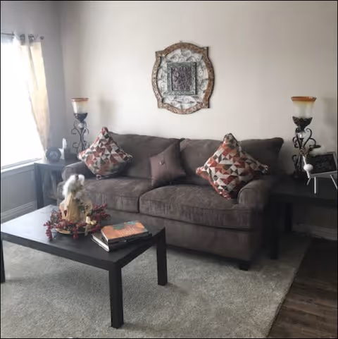 A cozy living room with a dark gray sofa adorned with three decorative pillows, two with geometric patterns and one solid brown. A black coffee table in front holds a decorative angel figurine with a wreath and two books. On either side of the sofa are matching dark side tables with ornate candle holders and lamps. A decorative framed wall art piece hangs above the sofa. Light filters in from a window with sheer curtains on the left side.