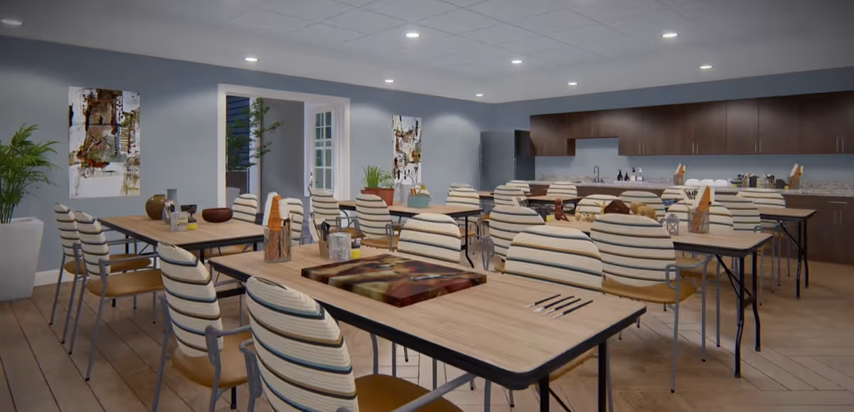 Large communal dining/activity room with many striped chairs and wooden tables and a kitchenette along the back wall.