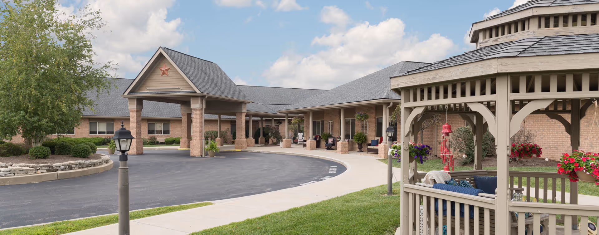 Exterior view of a senior living facility with a covered entrance supported by brick columns, a curved driveway, and a gazebo with seating and hanging flower baskets in the foreground. The building has a gray shingled roof and beige brick walls, with some greenery and trees around the area under a partly cloudy sky.