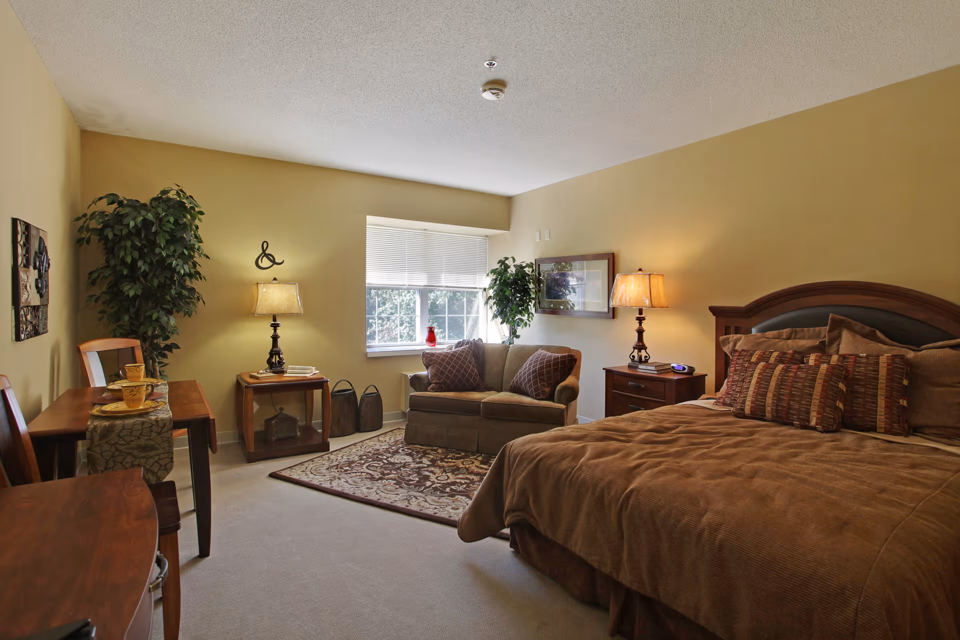 A cozy bedroom in a senior living facility with a large bed covered in brown bedding and multiple pillows. There is a wooden nightstand with a lamp and an alarm clock next to the bed. A small loveseat with two patterned pillows is placed near a window with blinds. The room also features a wooden table with two chairs, a decorative rug, two potted plants, and warm yellow walls with framed artwork.