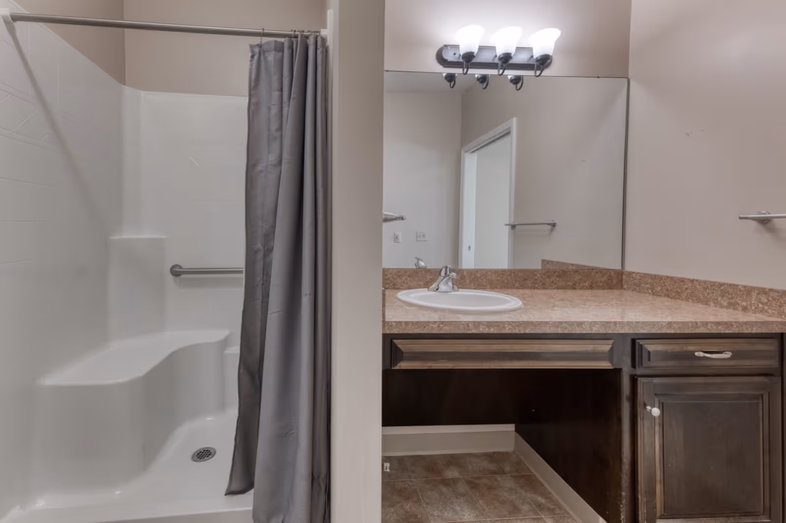 A bathroom with a white shower area featuring a built-in seat and a gray shower curtain. Adjacent to the shower is a vanity with a brown countertop, a white sink, dark wood cabinets, a large mirror, and a light fixture with three bulbs above the mirror.