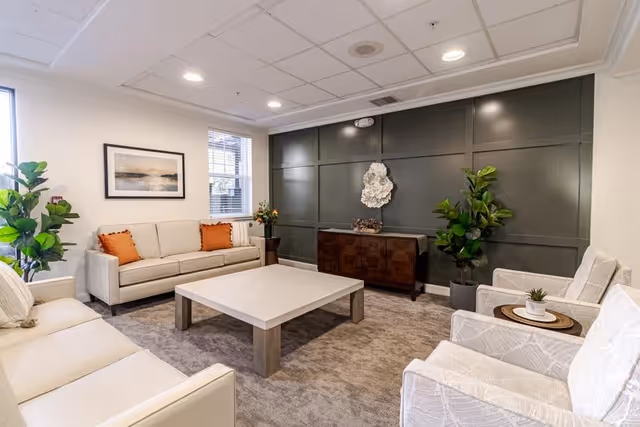 A cozy living room with two beige sofas, two patterned armchairs, a large square coffee table in the center, a dark accent wall with decorative art, potted plants, and a framed picture on a white wall near a window.