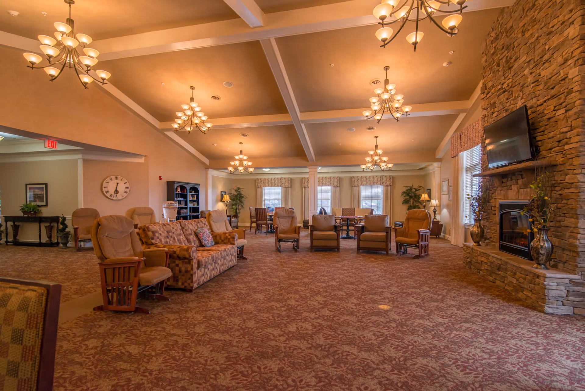 Spacious, warmly lit communal lounge with sofas and armchairs, chandeliers, and a stone fireplace with a mounted TV.