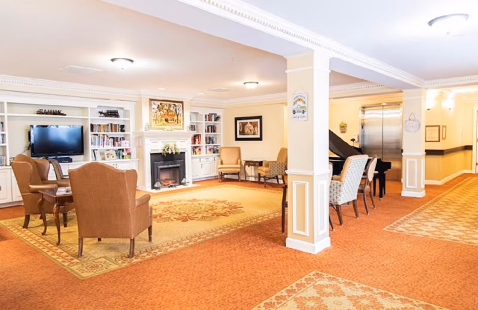 A spacious and well-lit common area in a senior living facility featuring comfortable armchairs arranged around a fireplace with built-in bookshelves and a flat-screen TV. The room has warm-toned carpeting with patterned rugs, decorative wall art, and a grand piano near an elevator in the background.
