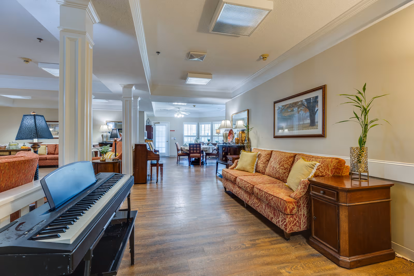 Interior view of a senior living facility common area with a patterned orange sofa, wooden side tables, a digital piano, and a dining area in the background. The room has wooden flooring, beige walls, framed artwork, and decorative plants.