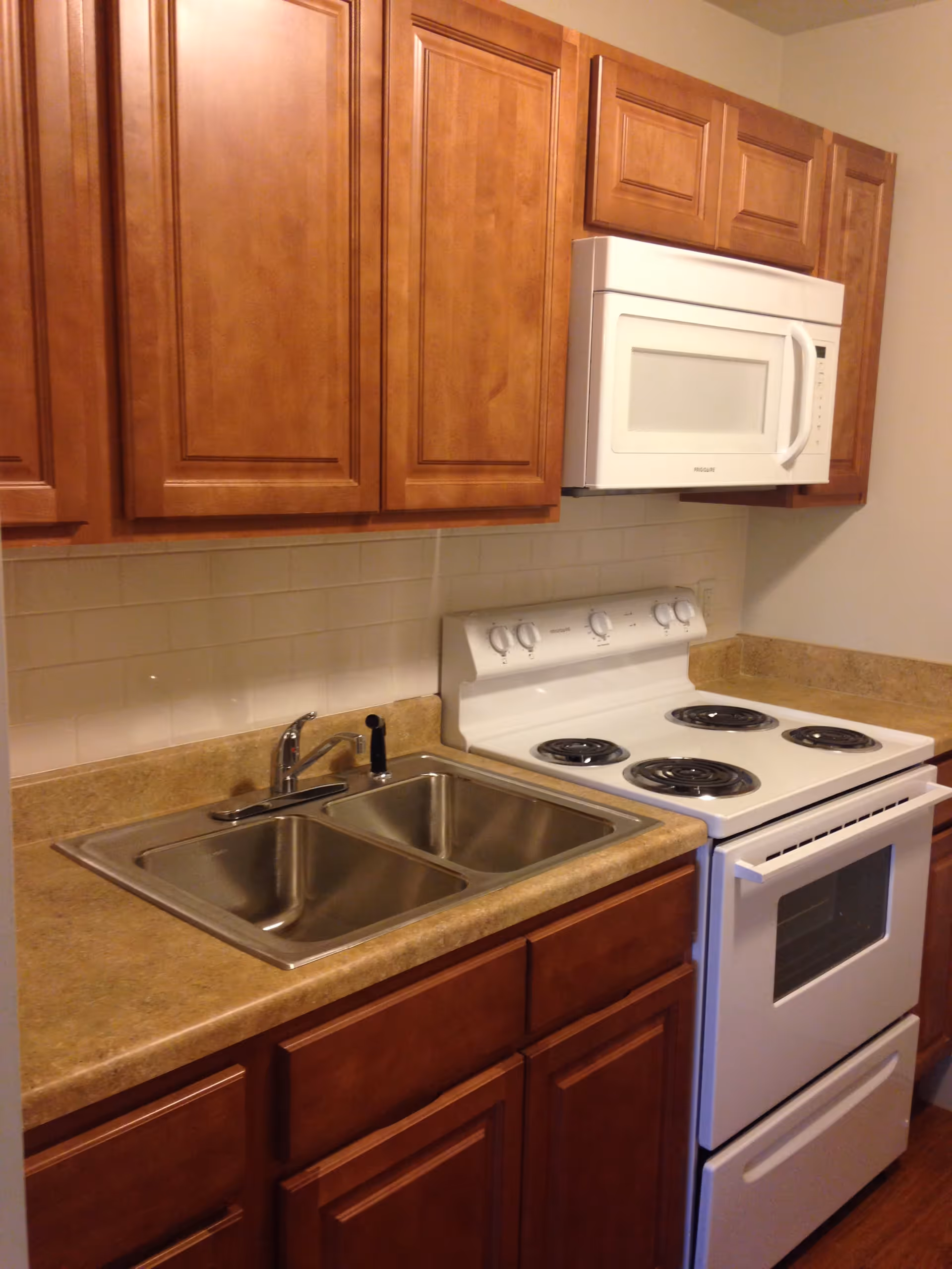 A kitchen area with wooden cabinets, a double stainless steel sink, a beige countertop, a white electric stove with four coil burners, and a white microwave mounted above the stove. The backsplash is made of white subway tiles.