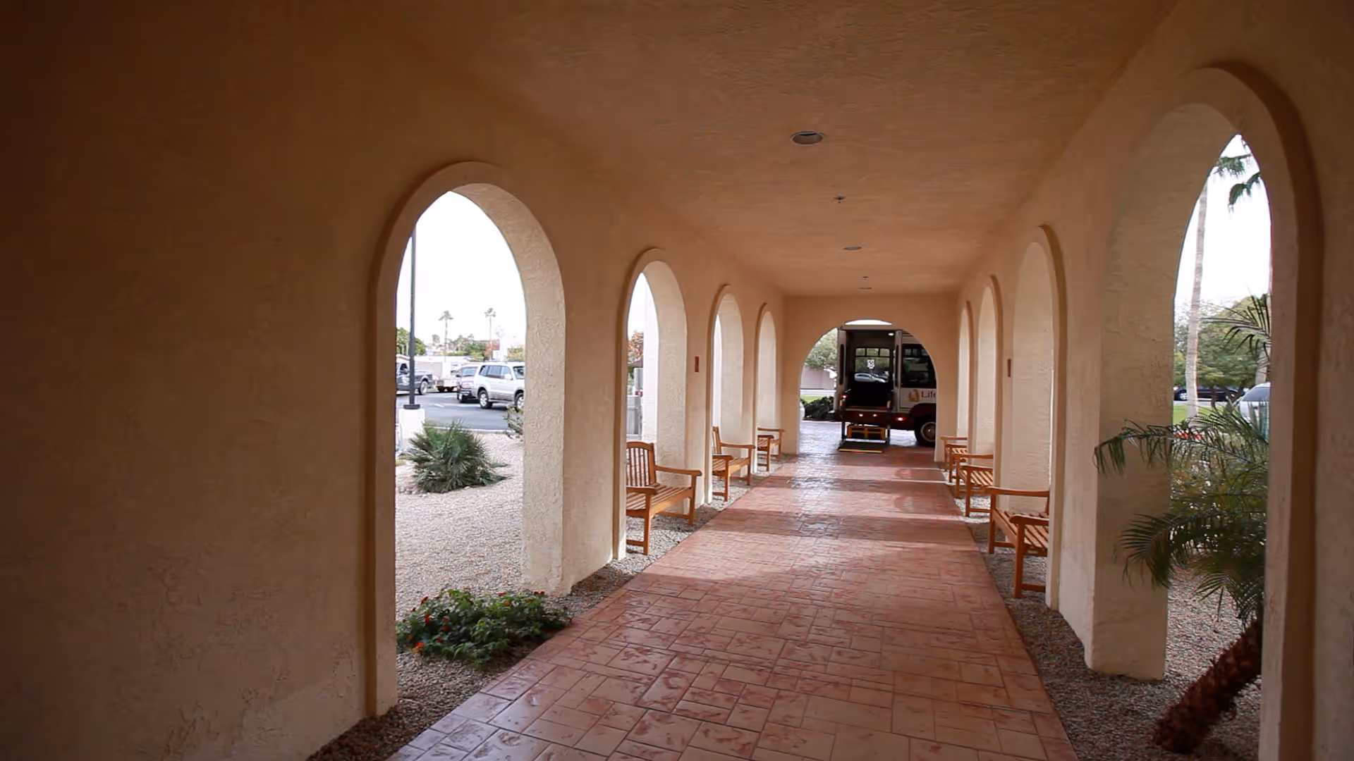 Covered outdoor walkway with arched openings on both sides, lined with wooden benches. The walkway has a tiled floor and leads to a parked shuttle bus at the far end. Desert landscaping with gravel and plants is visible outside the arches.