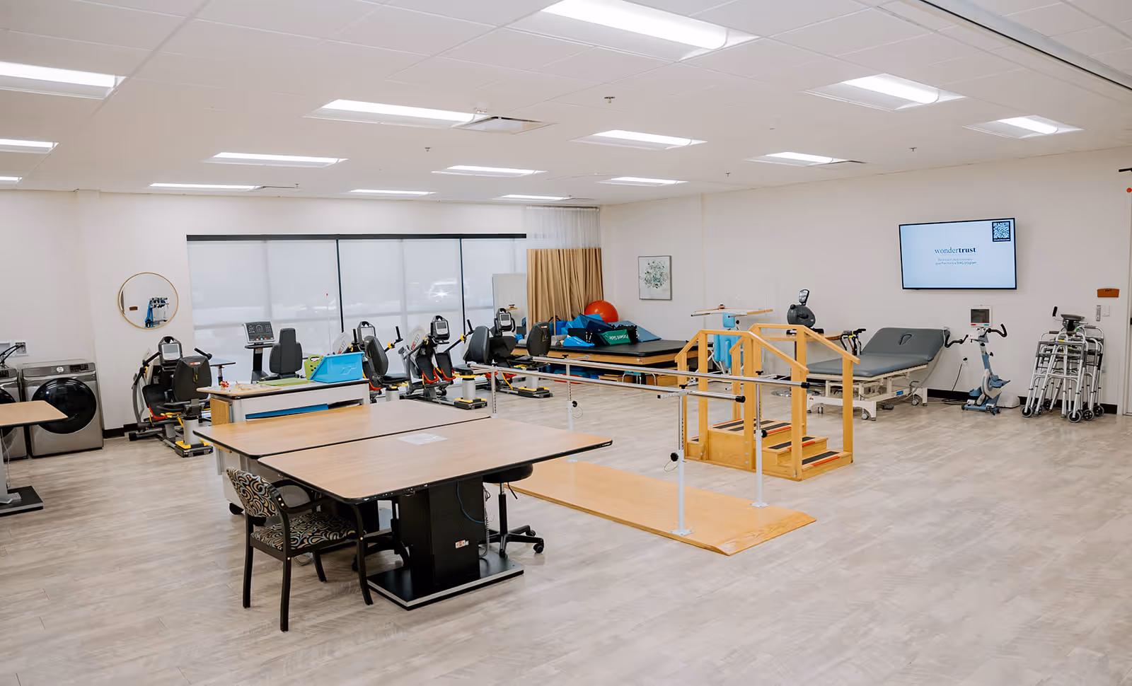 A spacious rehabilitation therapy room with exercise equipment including stationary bikes, parallel bars with wooden steps for walking practice, therapy tables, and various physical therapy tools. The room has large windows with blinds, light-colored flooring, and bright overhead lighting.