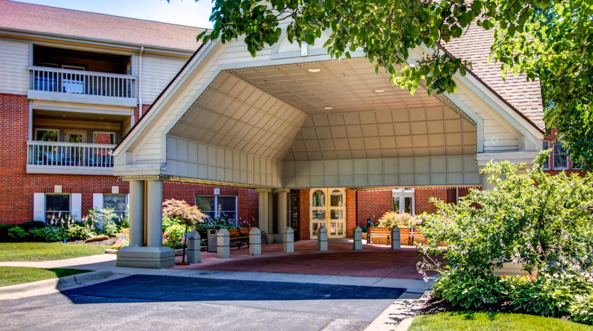 Entrance of a senior living facility with a covered drop-off area supported by columns. The building has red brick walls, white trim, and balconies. There are benches and landscaping with green bushes and trees around the entrance.