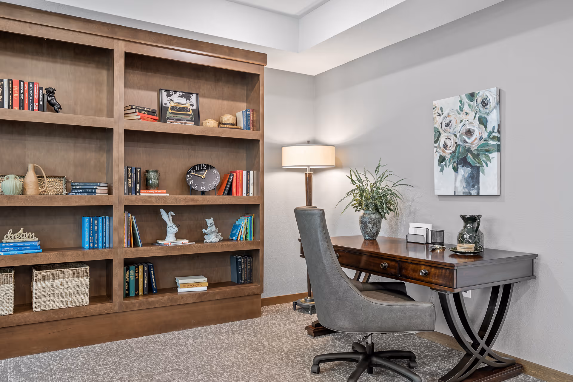 A cozy interior room featuring a wooden bookshelf filled with books, decorative items, and baskets. Next to the bookshelf is a wooden desk with a gray upholstered office chair. On the desk are a vase with green plants, a small organizer, and a decorative pitcher. A floor lamp with a beige shade stands behind the chair, and a floral painting hangs on the light gray wall.