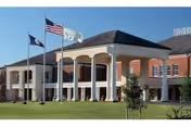 Exterior view of a large brick building with white columns and a covered entrance. Three flagpoles stand in front of the building, displaying the American flag, a state flag, and another flag. The building is surrounded by a well-maintained lawn and small trees under a clear blue sky.
