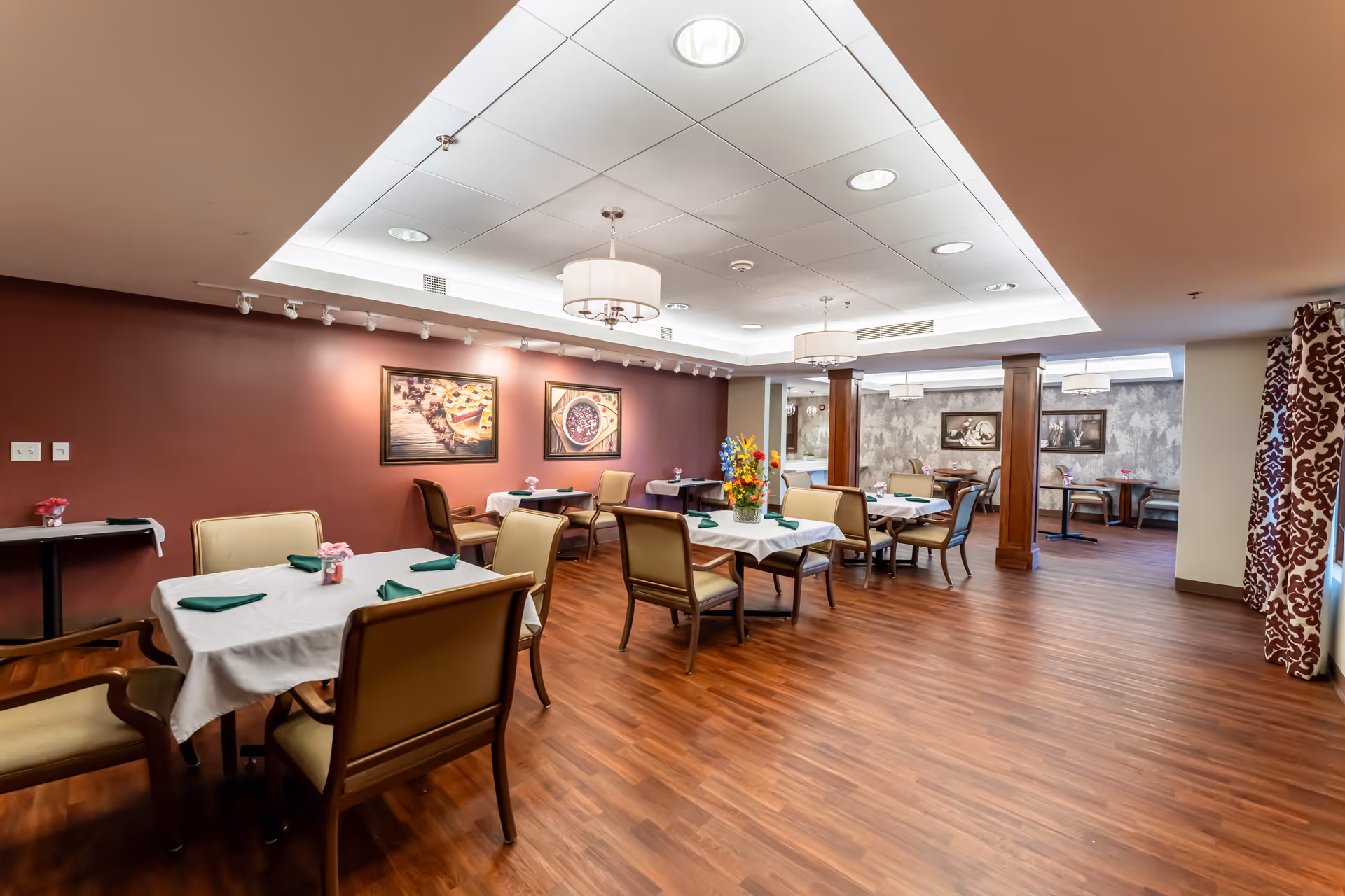 Dining room with several small tables set with napkins and chairs, hardwood floors, artwork on the walls, and ceiling lighting.