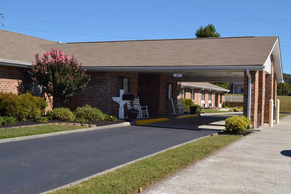 Covered brick building entrance with a drive-through portico, rocking chairs on the porch, and landscaped shrubs.