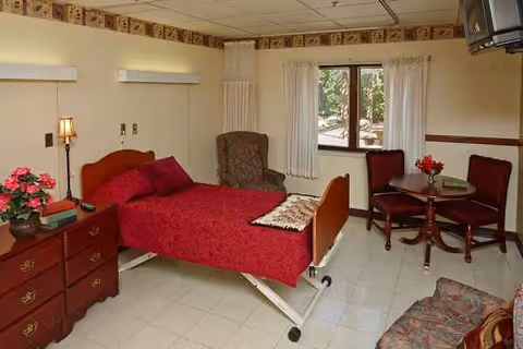 A cozy resident bedroom with a red bed, dresser, seating area, and window.