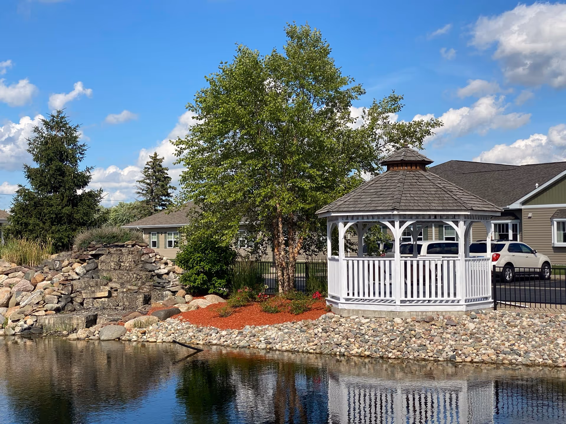 A white gazebo beside a pond with a rocky shoreline, trees, and nearby residential buildings under a blue sky.