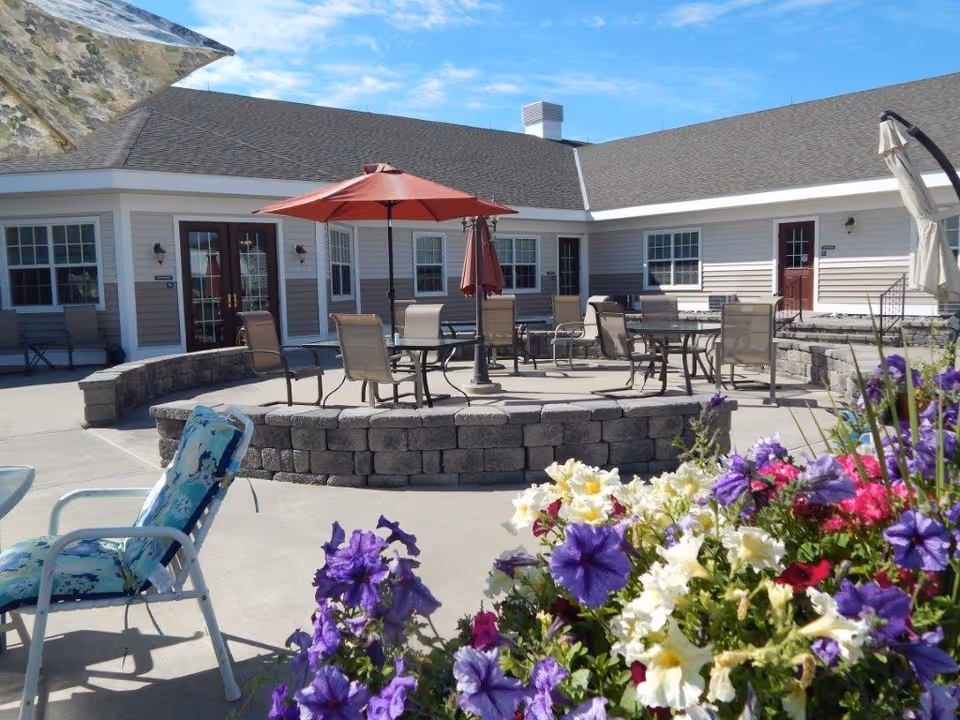 Outdoor courtyard with patio tables, chairs and umbrellas outside the assisted living building with colorful flowers in the foreground.