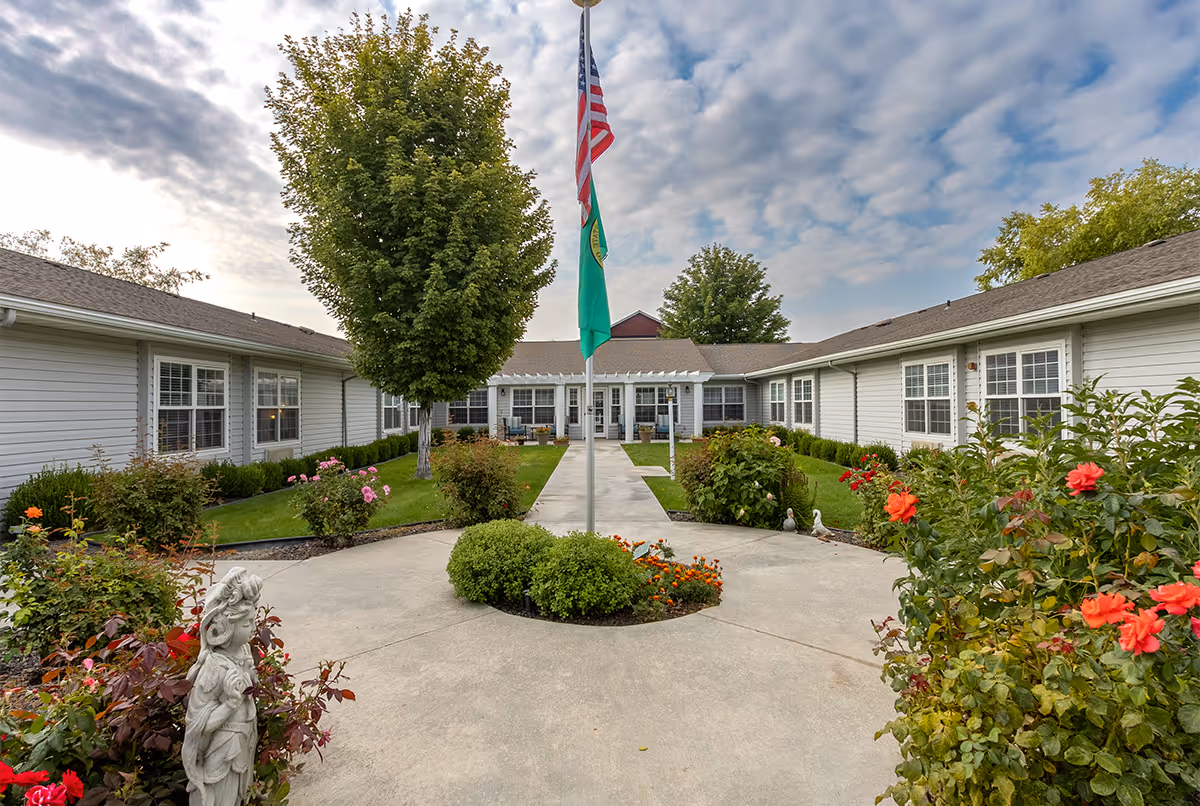 Outdoor courtyard area of Avista Senior Living Yakima featuring a concrete pathway leading to the building entrance, surrounded by green grass, bushes, colorful flowers, and a tree. Two flags are displayed on a flagpole in the center of the pathway under a partly cloudy sky.