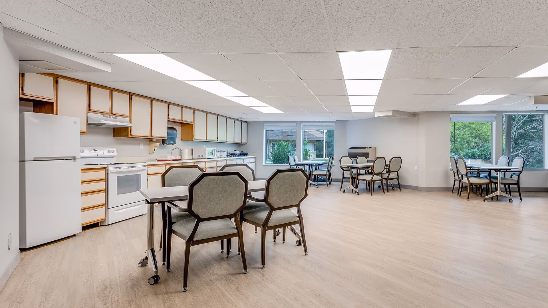 A spacious dining area with multiple tables and chairs arranged throughout the room. The room features a kitchen area along one wall with white appliances including a refrigerator, stove, and microwave, as well as wooden cabinets. Large windows allow natural light to enter, and the floor is covered with light-colored wood or laminate flooring. The ceiling has recessed lighting panels.
