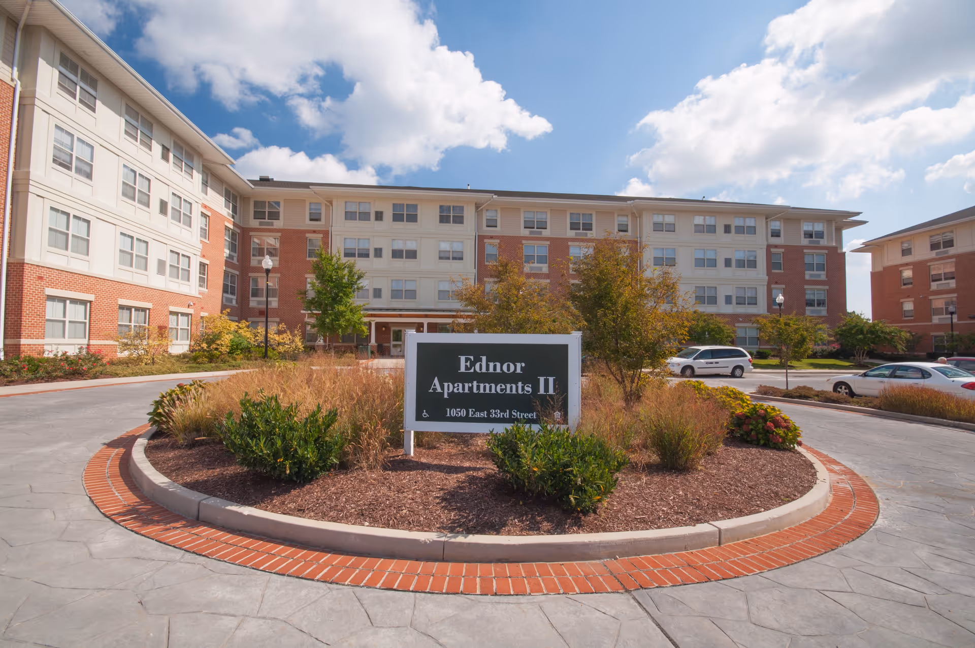 Exterior view of Ednor Apartments II, a multi-story residential building with red brick and white paneling. In front of the building is a circular landscaped area with bushes and a sign that reads 'Ednor Apartments II, 1050 East 33rd Street'. Several cars are parked around the building under a partly cloudy sky.