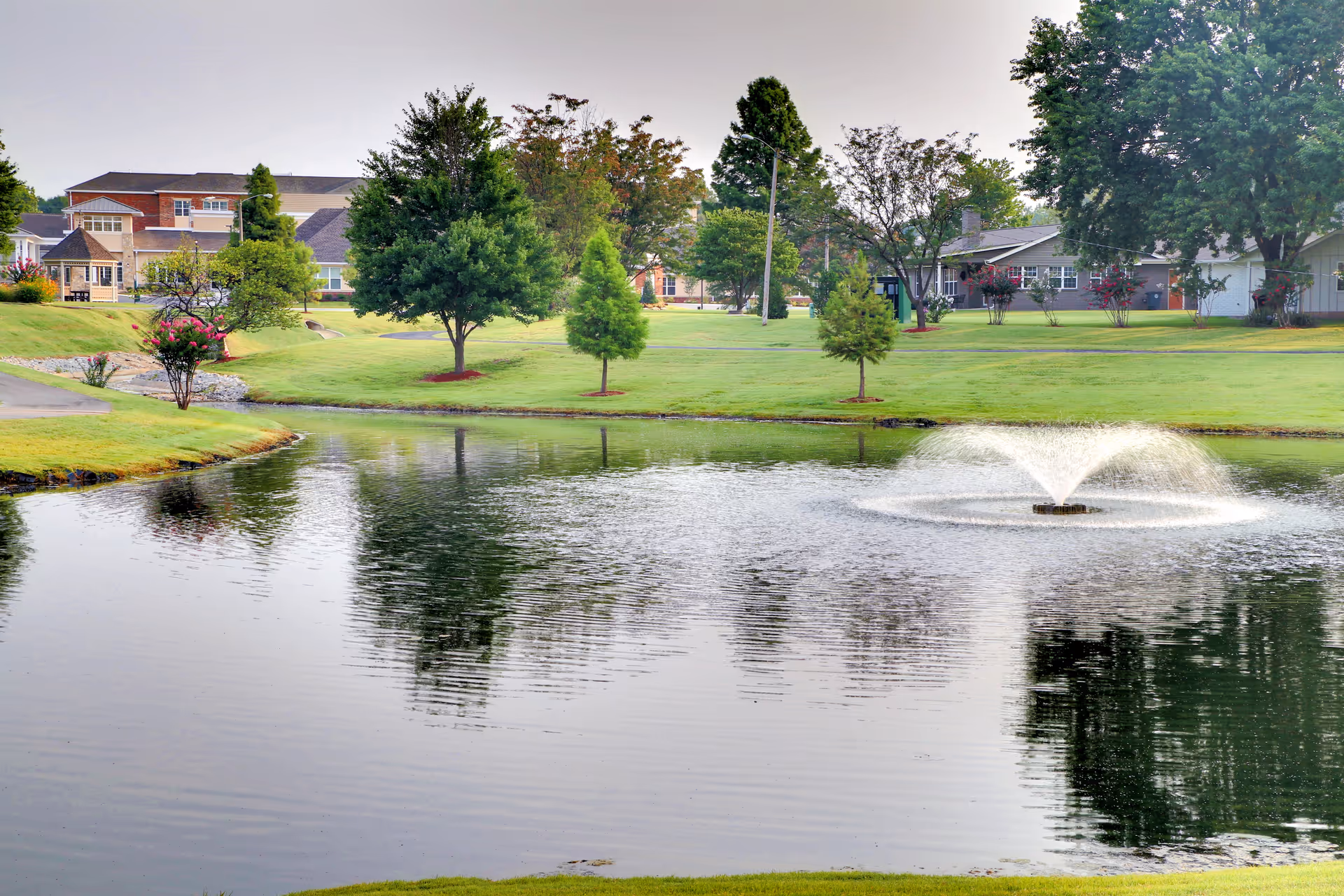 A pond with a central fountain surrounded by landscaped lawns, trees, and residential buildings in the background.