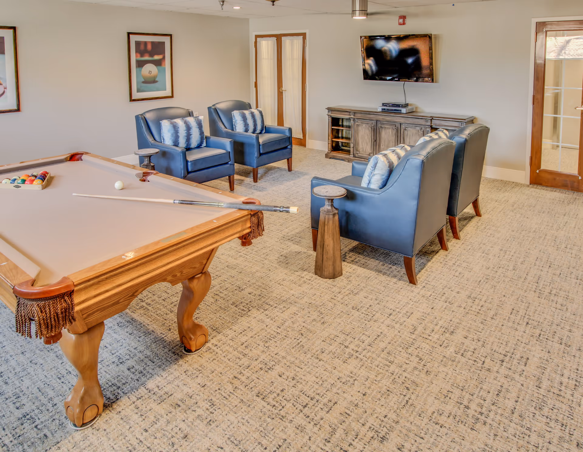 A recreational room featuring a pool table with a cue stick and balls, four blue armchairs with patterned cushions arranged around a small wooden side table, a wooden cabinet with a flat-screen TV mounted above it, and framed artwork on the walls. The room has carpeted flooring and glass doors.