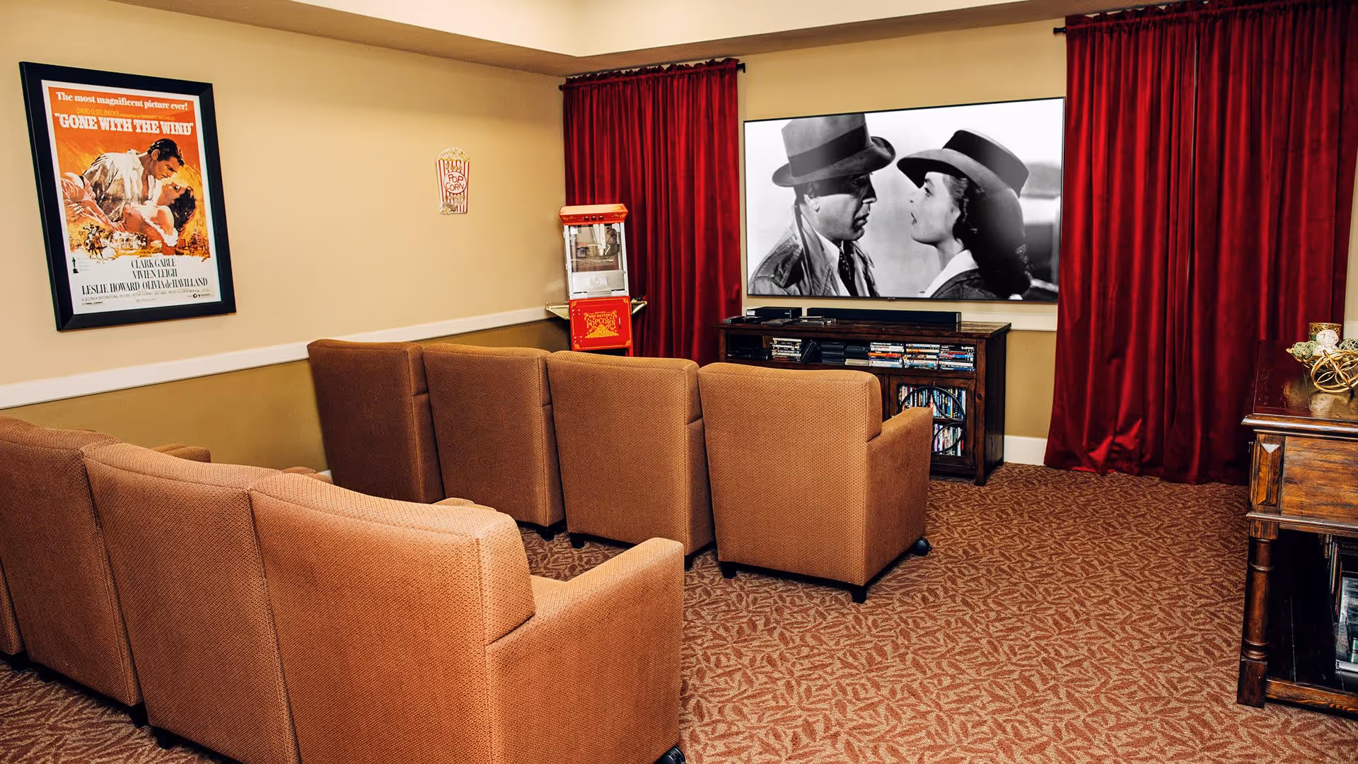 A cozy movie room with a row of brown upholstered chairs facing a large screen showing a black and white film. The room has red curtains on either side of the screen, a popcorn machine in the corner, a framed Gone With The Wind movie poster on the wall, and a wooden cabinet holding DVDs and media equipment.