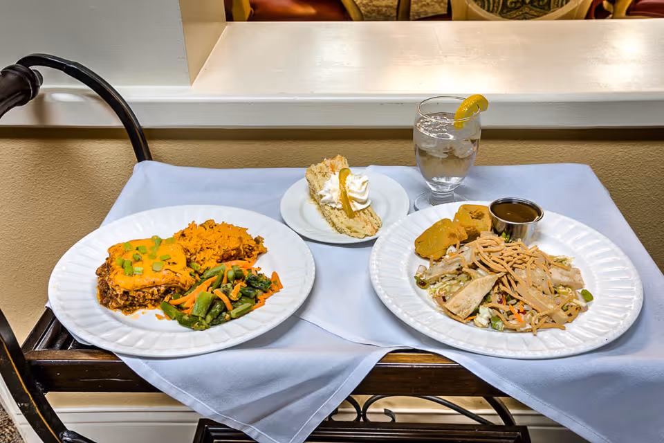 A meal served on a table with a white tablecloth, featuring two plates of food, a slice of cake with whipped cream and a lemon garnish, and a glass of water with a lemon wedge. One plate has a serving of lasagna, rice, and mixed vegetables, while the other plate has a salad with chicken, crispy noodles, a small bread roll, and a cup of dressing.