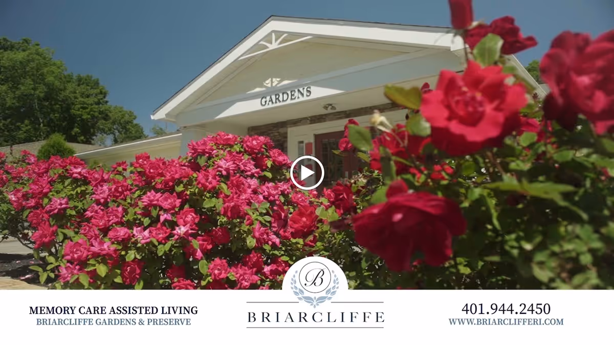 Entrance of Briarcliffe Gardens Memory Care Assisted Living facility with vibrant pink flowers in the foreground and a clear blue sky above.